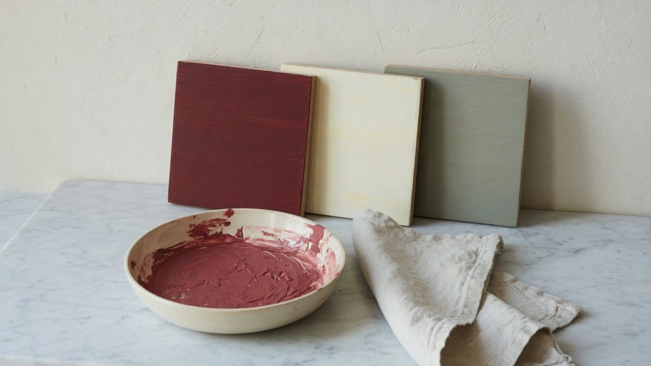 Milk paint powder, mixing bowl, and sample boards in muted historic reds and creams in a MetroWest workshop, natural light